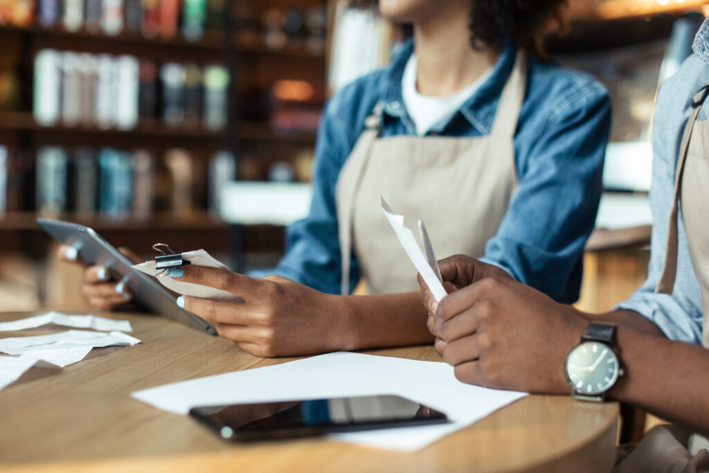 Cafe shop, small business, accounting and bill payment. Focus on hands of african american millennial male and female, owners with tablet and papers in hands on table with smartphone, cropped