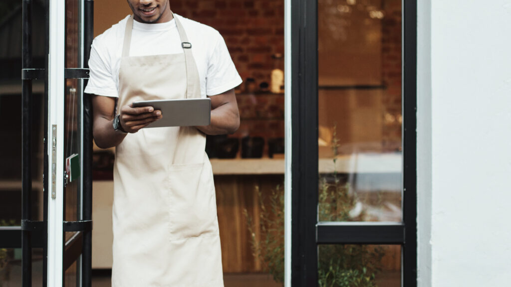New normal and ordering coffee online for takeaway during covid-19 quarantine. Millennial african american male in apron works on tablet near front door of modern cafe, cropped, empty space