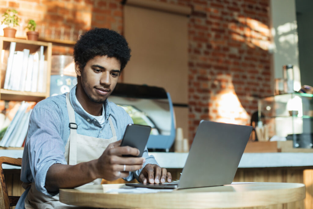 Busy african american man in apron sits at table and works in smartphone and laptop 